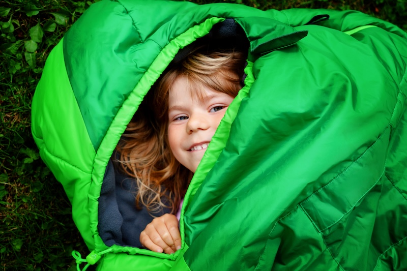 Preschool little girl in sleeping bag camping. Outdoors activity with children in summer. Fun and adventure camp, family and friends vacations or weekend trip. Portrait of child with flashlight.